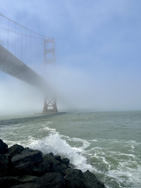 The "International Ornage" Golden Gate Bridge on the left, shrouded in fog, with fading blue skies above. The mists thickened and waned while we were watching. Green bay waters splash across dark rocks in the lower left.