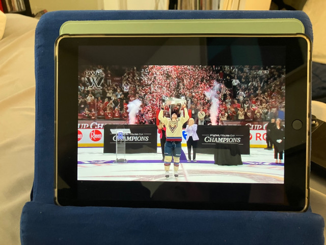A tablet in a thick blue protective case rests on a bed or soft surface indoors. On the tablet screen, a hockey championship celebration is playing. A player in a cream-colored jersey stands at center ice holding a large silver trophy overhead while red confetti explodes behind them. Smoke effects rise on both sides of the stage area, and a packed arena crowd fills the background. “PWHL Walter Cup Champions” branding is visible around the rink boards and stage setup. The room around the tablet is softly lit, with shelves and household furniture blurred in the background.