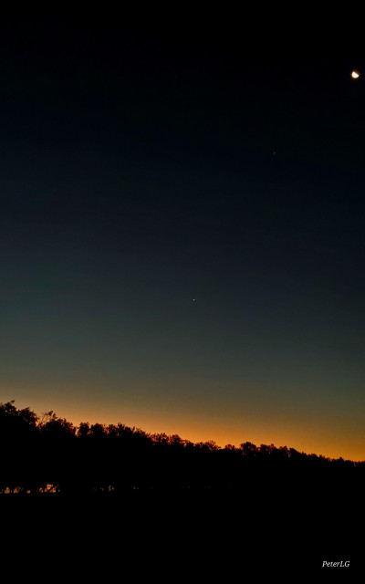 Sunset picture, with the sky graduating from black to deep orange, the treeline appearing as black shadows. At the lower left can be seen a sliver of gold from the sky reflecting off of the river. At the top right can be seen the crescent moon. In the middle, if one looks c,osely, can be seen Venus.