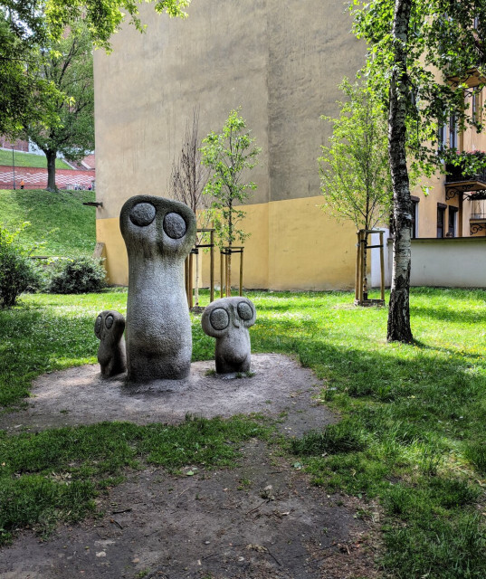 A serene park scene in Kraków featuring three whimsical, abstract stone owl sculptures by Bronisław Chromy. The central owl stands tall with wide, expressive eyes, flanked by two smaller owls in a playful arrangement. The sculptures are set on a patch of dirt surrounded by lush green grass, young trees, and a yellow building in the background.