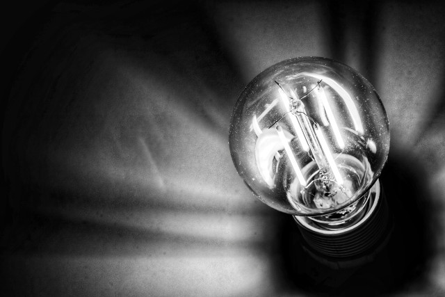 Das Schwarz-Weiß-Foto zeigt eine Nahaufnahme einer brennenden LED-Filament-Glühbirne in einer dunklen Umgebung. Die Glühbirne befindet sich in der rechten Bildhälfte und strahlt ein helles, kühles Licht aus, das die feinen vertikalen Leuchtfäden im Inneren des Glaskolbens deutlich sichtbar macht. Von der Lichtquelle ausgehend werfen die Filamente strahlenförmige, weiche Schatten nach links oben und unten auf eine strukturierte, stoffähnliche Oberfläche im Hintergrund. Der untere Teil der Birne verschwindet in einer tiefschwarzen Fassung, während der Rest des Bildes durch ein kontrastreiches Spiel aus hellem Licht und tiefen Schatten geprägt ist, was der Szene eine dramatische und fokussierte Atmosphäre verleiht.