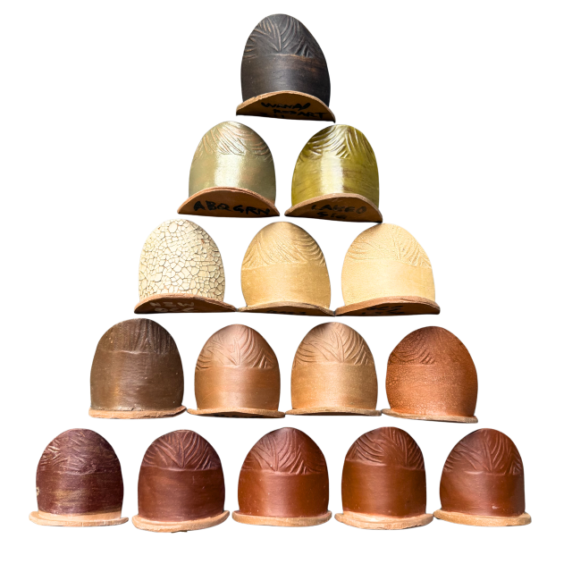 A pyramid of small ceramic test tiles, each rounded on top and flat on the bottom. The top tile is black; the second row of two are tones of olive green; the third row of three are tones of off-white and yellow (some with crackle texture); the fourth row of four are tones of brown, tan and orange; the final row of five are variations of dark iron red.