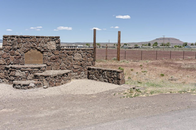 A color landscape photo of a monument made of large brown colored stones on the left. It has places to sit in front and a metal plaque on a wall in the back. To the right is a gate bordered by tall wood posts and a fence dividing the short weeds in front and a reddish gravel surface behind the fence. There are a few trees in the distance and a dome shaped hill in the background on the right. The sky is mostly clear, blue, with six small clouds.