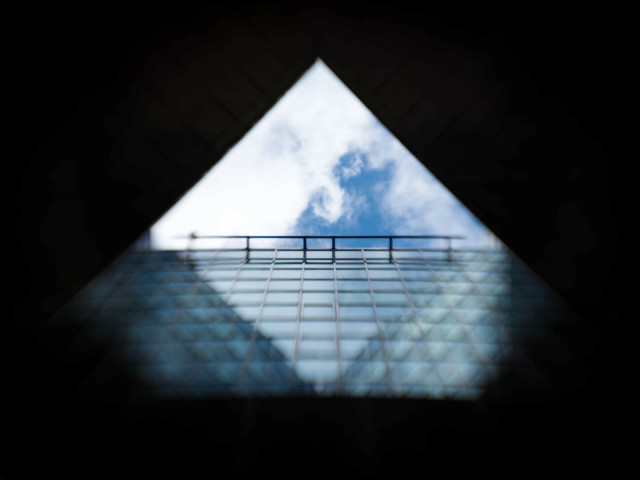 Looking up at a triangle of blue sky and white cloud, hemmed-in by darkness on the top two sids, and on the bottom by a wall of gridded glass. In the windows both the sky and the other sides of the triangle are reflected, showing that the latter are also all glass panes.The photo begins to blur outside of a central in-focus disc.
