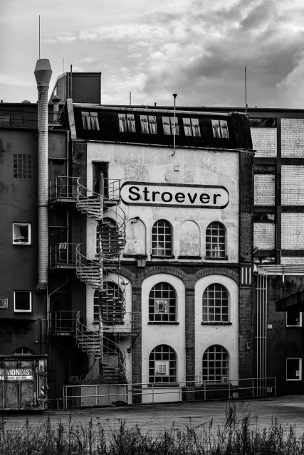 Ein Industriegebäude in Schwarz-Weiß mit einem auffälligen Schild, auf dem „Stroever“ steht. Es verfügt über eine spiralförmige Feuertreppe an der Außenseite, große Bogenfenster und eine facettenreiche Fassade. Der Himmel ist bewölkt, was zur
