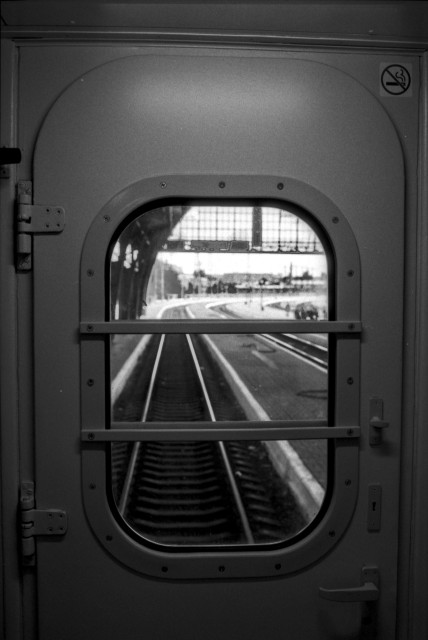 Black-and-white film photograph of the rear door of a train car, with an oval window framed by two horizontal safety bars. Through the window, railway tracks recede into the distance inside a large station, with a platform and overhead structure visible. The door features a handle, hinges, and a no-smoking sign in the upper right corner. Shot on Ilford FP4.