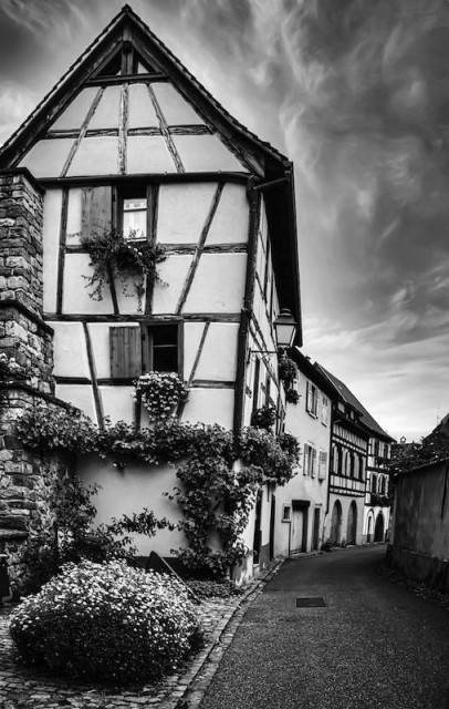 Man sieht eine schmale Gasse, wo vielleicht ein Auto durchfahren kann. An der linke Seite sieht schmale, mehrstöckige Fachwerkhäuser die bis zur Kante der Gasse reichen. Am Himmel sieht man Schleierwolken. Das Foto ist in Schwarzweiß.