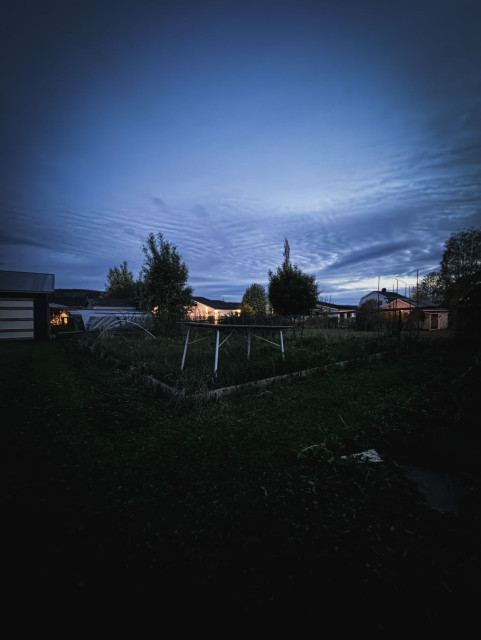 A dark suburban garden at blue hour, bathed in deep blue twilight. A white metal table stands among overgrown raised beds and patches of grass. Trees silhouette against the pale sky and warm lights from neighbouring houses glow in the background. Wispy clouds streak across the blue-grey sky above. The scene is quiet, slightly melancholic and deeply still.