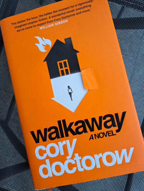 A picture of "walkaway" a novel by Cory Doctorow, here in its first hardcover edition. The dustjacket is cut (by design), and slightly torn on the cut area. The cover is bright orange with a black house on fire.