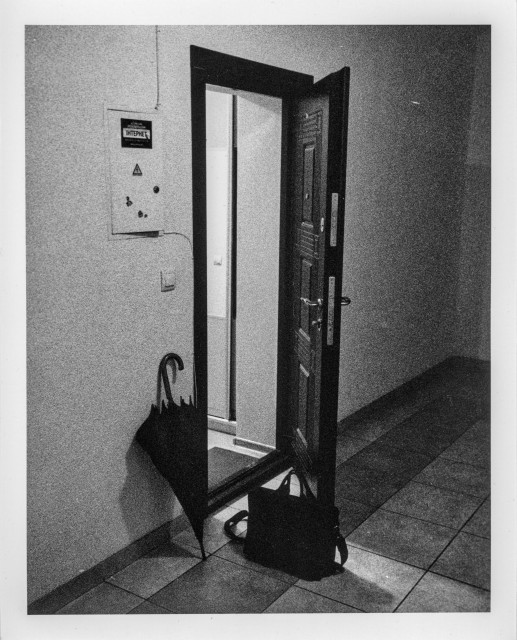 A grainy black-and-white photograph of a quiet hallway. An open door reveals a brightly lit interior space, creating a stark contrast with the darker corridor outside. Near the doorway, a closed umbrella leans against the wall, while a dark bag rests on the floor beside the threshold. On the wall, there is a small electrical panel labeled “Internet,” adding an unexpected mundane detail to the otherwise cinematic scene. The strong contrasts, rough film texture, and empty space create a feeling of solitude, tension, and quiet anticipation — as if someone has just arrived or silently left moments before.