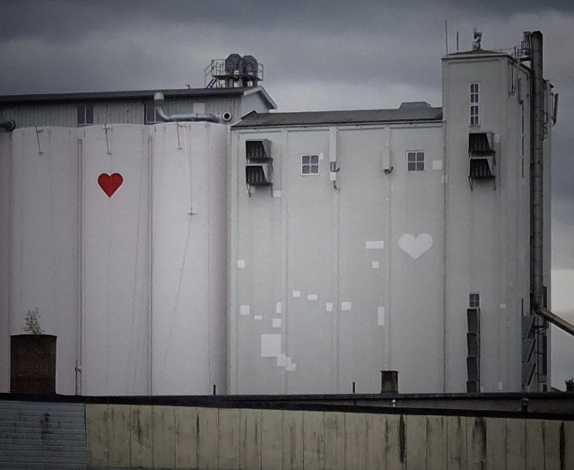 Grey and white silos in an industrial area where a rather small red heart is painted on one,  and a white heart on another.