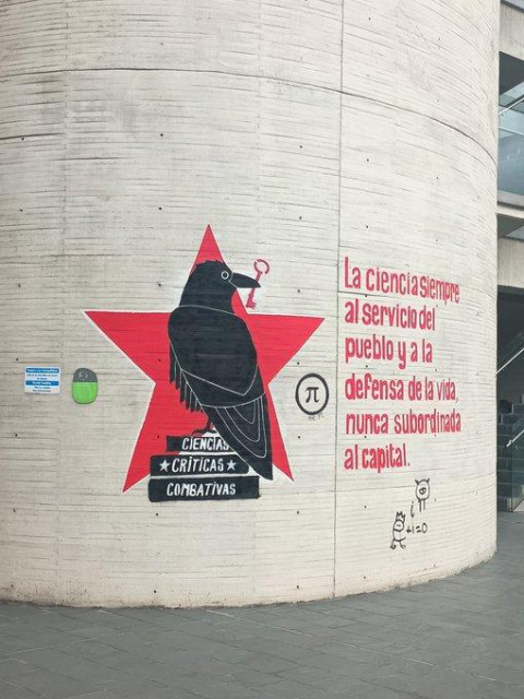 Image shows a white wall.  A large red star with a black bird holding a key, with the accompanying text "la ciencia siempre al servicio del pueblo y a la defensa de la vida, nunca subordinada al capital".
