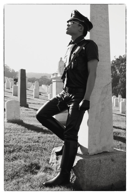A monochrome picture of a 20-something man fully decked out in leather (boots, pants, shirt, tie, gloves). He is gazing upward while leaning against an obelisk-shaped grave marker in a cemetery on a bright but hazy day.