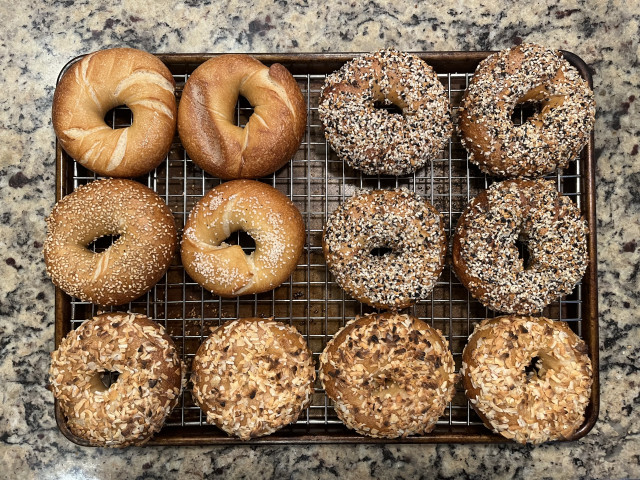 A dozen bagels cooling on a wire rack set in a baking sheet. At top left there are two plain bagels side by side, with a long scorch mark visible on the left side of the right hand bagel. Below those two bagels topped with sesame seeds (left) and salt (right). At top right there are four bagels coated with everything seasoning. On the bottom is a row of four onion bagels.