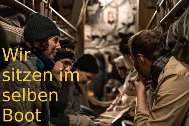 Das Innere eine U-Boots. Personen sitzen links und rechts auf Kojen und unterhalten sich, offenbar gut gelaunt. Sie tragen beide Uniformen oder Jacken, dazu Strickmützen. Text am Rand: "Wir sitzen im selben Boot"