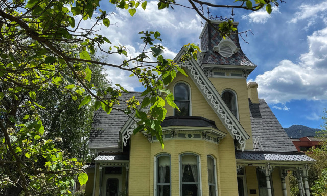 An historic home from the 1800’s with angled roof pitches, big windows, a cupola, bright yellow siding, and slate roof tiles, partially hidden behind green trees branches in bright afternoon sunshine and partly cloudy skies.