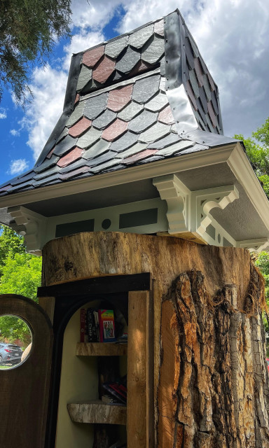 The large stump of a former cottonwood tree has been hollowed out inside to contain a small, free library with book-filled shelves behind a full-sized wooden door, which is open to see inside the stump. A hint of partly cloudy blue skies is visible above the cupola roof of the tree stump.