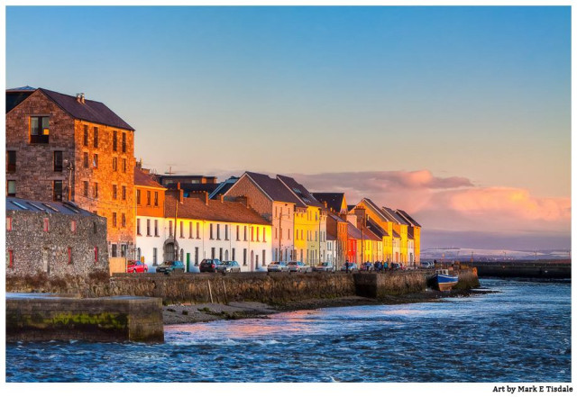 Color photograph of a row of buildings hugging the waterside on the Irish coast. The warm light and the skies around them suggest sunset is occurring out of frame. 