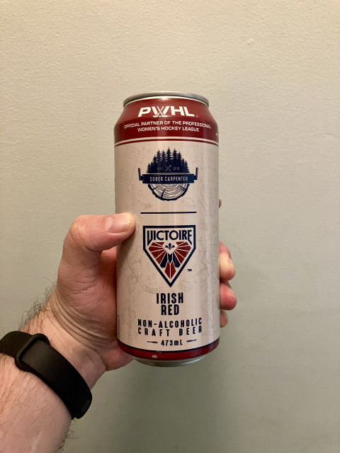 A hand holds a tall 473 mL can of non-alcoholic craft beer against a plain beige wall. The can is branded for the PWHL and features a collaboration between Sober Carpenter and the Montréal Victoire women’s hockey team. The design uses red, white, and navy blue colours, with the Victoire crest prominently centered above the words “Irish Red.” The top rim of the can reads “Official Partner of the Professional Women’s Hockey League.” The person holding the can wears a black fitness band on their wrist.