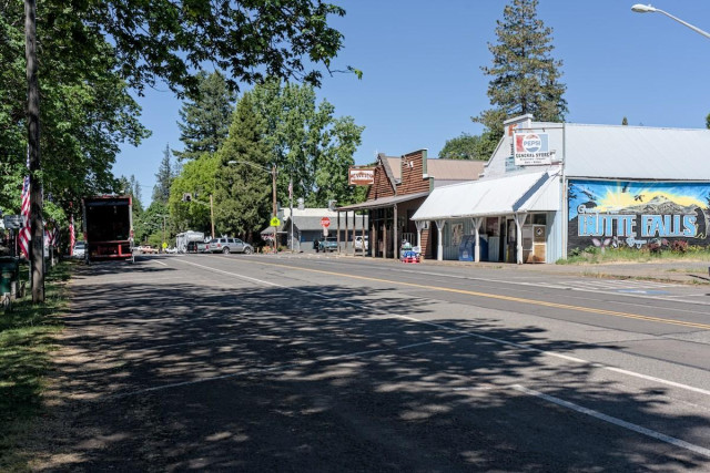 A color landscape photo looking down an empty street in a small town. On the right is a general store with a mural on the side that reads, " Butte Falls, Oregon." It shows a forest covered mountain with golden light, a blue sky, and birds flying around. Next to the general store and its covered porch is a log building that has a sign that says, "Tavern." There's a stop sign and then a couple more buildings. Conifer trees are seen in the background. The photo is taken from the left side of the street and along the left edge of the frame there is a line of trees with US flags vertically mounted between them. 