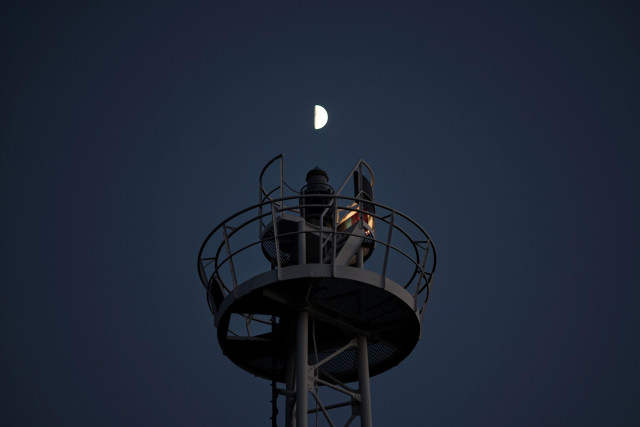 Oberer Teil eines Leuchtturms aus Metallstreben bei Nacht. An der rechten Seite erkennt man das Leuchtfeuer, genau über der Spitze des Turmes steht der Halbmond. 