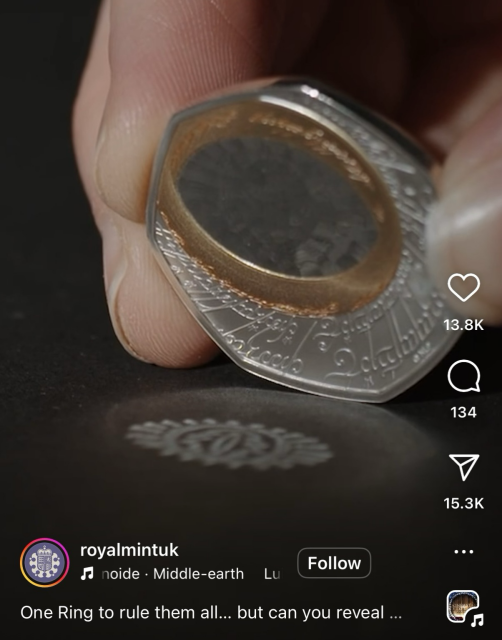 Closeup of a hand holding a 50p coin. The coin is silver with engraved script, and an off centre gold coloured detail for ‘the one ring’ from The Lord of the Rings. The middle of that ring area is polished and reflects The Eye of Sauron onto the table.