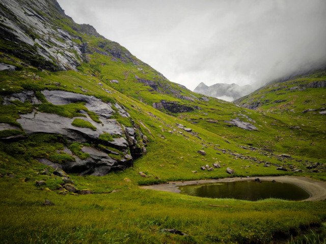 The image depicts a serene mountainous landscape with lush green grass covering the slopes. The foreground features a small, circular pond with dark water, surrounded by vibrant green grass and scattered rocks. The midground shows steep, rocky hillsides with patches of grass and moss, leading up to a misty background where the peaks of distant mountains are partially obscured by low-hanging clouds. The sky is overcast, with gray clouds dominating the scene, suggesting a cool and damp environment. The overall color palette is dominated by various shades of green, with the dark gray of the rocks and the muted tones of the sky providing contrast.