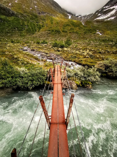 Das Bild zeigt einen roten Seilbrücken, der über ein wildes, fließendes Gewässer führt. Die Brücke ist aus Metall und hat eine schmale, flache Plattform mit Seilen, die sie an den Seiten festhalten. Unter der Brücke fließt ein türkisfarbenes, schnell dahinströmendes Wasser, das von einem felsigen Ufer umgeben ist. Auf beiden Seiten des Flusses wachsen grüne Büsche und Sträucher, und im Hintergrund erheben sich steile, grüne Hügel mit vereinzelten Schneeflecken. Der Himmel ist bedeckt, was auf eine bewölkte Atmosphäre hinweist. Die Landschaft wirkt wild und unberührt, mit einer natürlichen Schönheit, die durch die Brücke und das fließende Wasser betont wird.