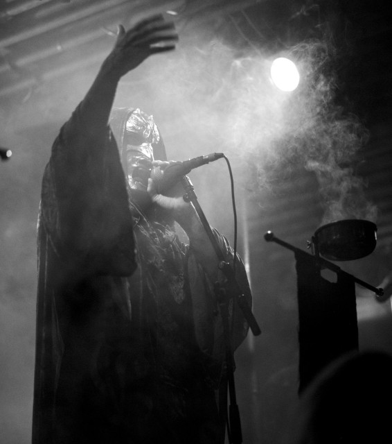 Schwarz-weiße Aufnahme eines Blackmetal-Sängers mit Kapuzenumhang und Mikrofon vor Nebel und Bühnenlicht