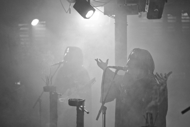 Schwarz-weiße Aufnahme eines Blackmetal-Konzerts mit zwei Musikern, von denen einer singend die Hände erhoben vor einem Mikrofon steht und der andere im Hintergrund Gitarre spielt, umgeben von Bühnennebel und Scheinwerfern.