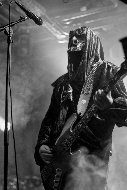 Musiker mit Kapuzenpullover und Maske spielt E-Bass auf einer verrauchten Bühne bei einem Blackmetal-Konzert