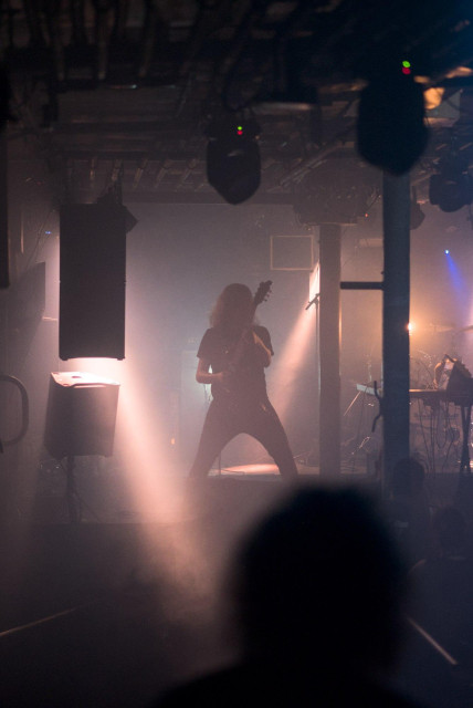 Silhouette eines Gitarristen mit gespreizten Beinen auf einer Bühne bei einem Blackmetal-Konzert, umgeben von Nebel und Bühnenlicht