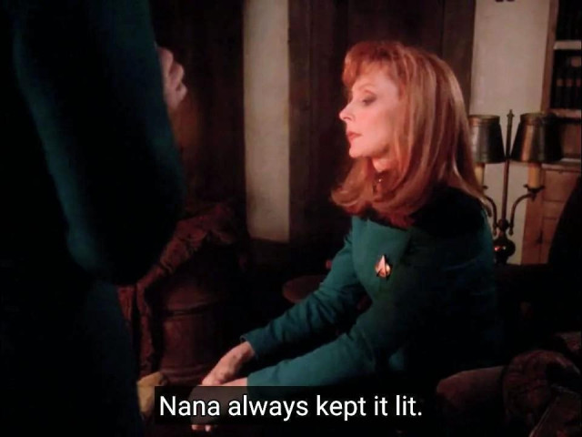 Star Trek The Next Generation scene. We're in an older earth looking home with some antiquey lamps in the background and a bookcase. From the center, sitting with hands on lamp looking wistfully down as if lost and thought is Dr Beverly Crusher. She's in her blue uniform which here looks teal but we know. It's blue. There's another standing near her but mostly off camera. Closed caption reads, "Nana always kept it lit."