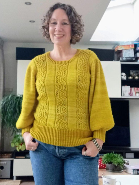 A woman with short curly hair is wearing a bright yellow sweater. She is smiling into the camera.