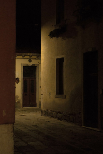 At night, a path zigzags under pale yellow light between doors set in pale plastered walls.