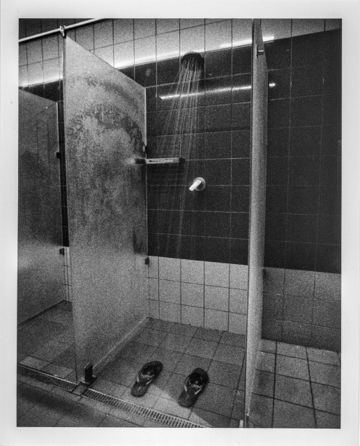 A grainy black-and-white photograph of an empty public shower room. Water pours from a large overhead showerhead onto tiled floors, while fogged glass partitions catch the light and create soft, ghostly textures. A pair of dark sandals rests beneath the running shower, suggesting someone has just left or is about to return. The scene feels quiet, intimate, and slightly liminal — a mix of solitude, routine, and cinematic atmosphere enhanced by the high contrast and visible film grain.