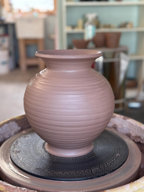 A freshly wheel-thrown vase on the potter’s wheel. The clay is light brown and has a soft sheen of wetness. It is round with a wide, flared neck. Blurred in the background: a shelf, a small kiln, a sink with a window above it. 