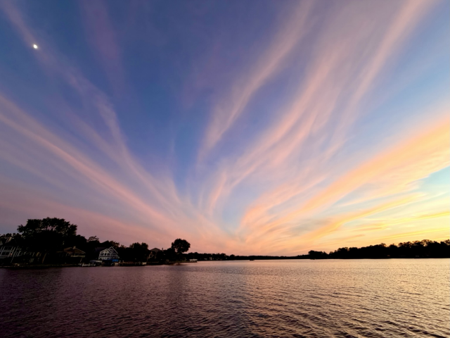 Sunset photo of pink clouds streaming up from the horizon over a lake with the Moon in the upper left.