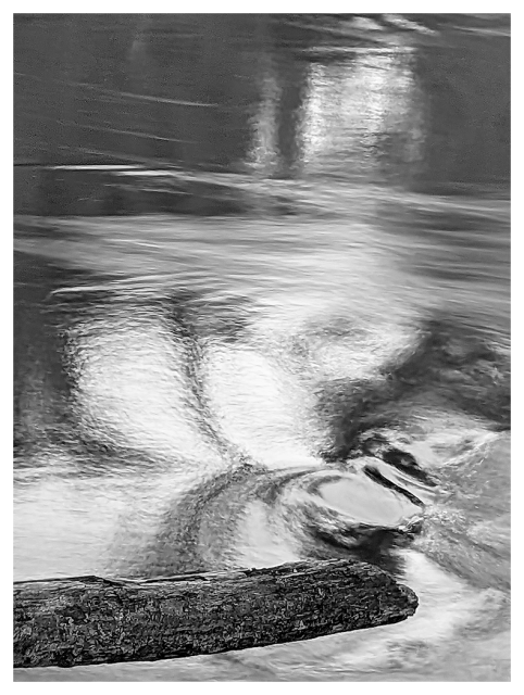 black and white photo of a swirling water surface, reflecting light in soft patterns. a rough, horizontal log lies across the lower part of the image, partially submerged, with ripples and eddies swirling around it.
