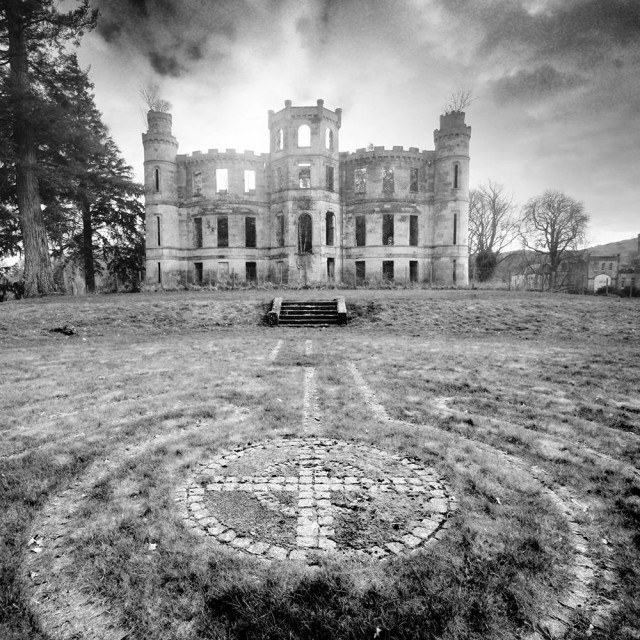 Black and white photo of a ruined Georgian style castle, surrounded by trees under dark clouds, with a brick maze laid in the lawn in the foreground.