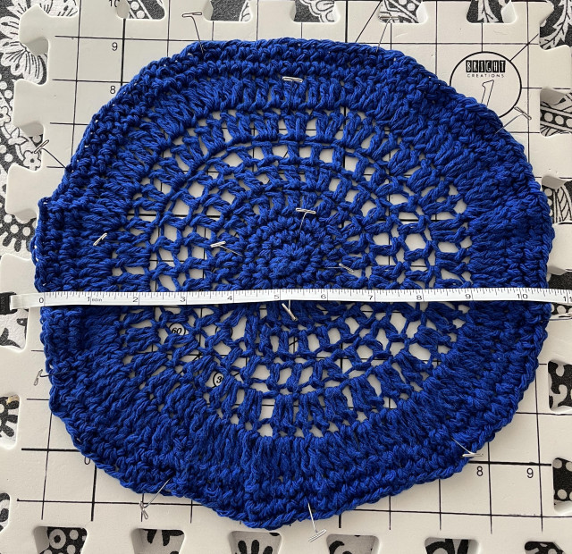 A badly crocheted round thing made from royal blue colored cotton yarn. It is a bit wider than 10 inches in diameter, as shown by the tape measure lying across it.