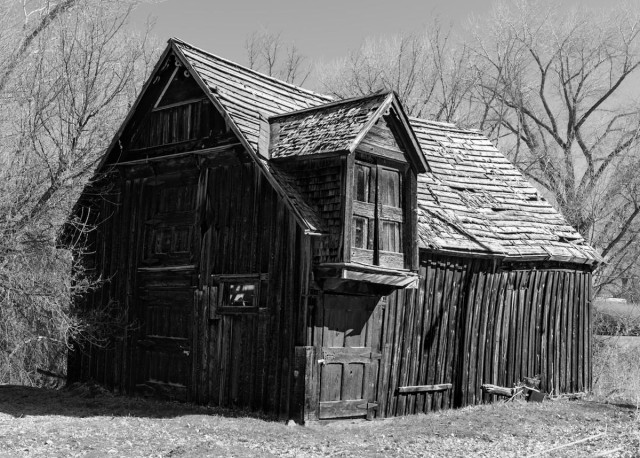 A black and white landscape image of an old weathered barn. The front is in shadow on the left. The right side is in daylight. There are two doors, one above the other. The lower door is a large single door. The upper door is two doors. They are set in a part of the barn that extends out from the main structure, but there is no landing. These doors accessed the upper part of the barn for loading hay bails. Winter bare trees surround the bar. 