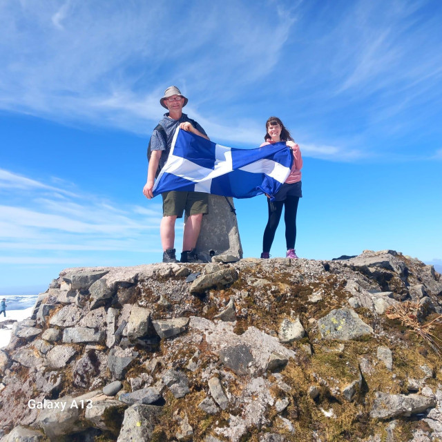 A photo of a middle aged man and his grown-up daughter standing atop the cairn at the summit of Ben Nevis (1,345 m, 4413 ft) on a lovely day in late spring. The sky is a beautiful blue with high-level wispy clouds, and the fool is making her hold up a Scottish Saltire between them 🏴󠁧󠁢󠁳󠁣󠁴󠁿😁