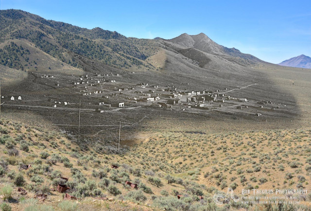 A color landscape photo composition. Two photos were used to make a "then and now" photo of a ghost town called "Wonder" in central Nevada. The original photo was made around 1911. It shows the small mining town on a gentle slope at the base of a mountain. That photo is in black and white. I found the spot where the photographer stood and shot the scene again using the same photographic settings. Then at home, I composited or blended the two photos such that the town, in black and white, is in the middle of the frame and extends up the side of one of the mountains and the rest is in color showing what the site looks like now. There are a few old rusty cans in the foreground. The sky is clear and blue.