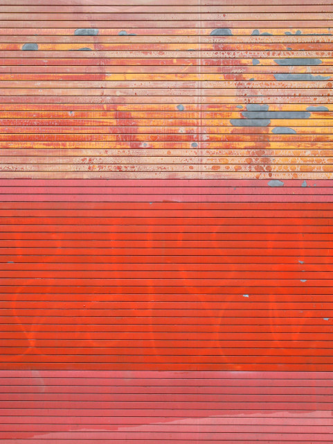 Photo of a metal roll-up door with very thin horizontal slats. The bottom two thirds is various shades of red, with some graffiti visible behind the darkest and newest layer of paint in the middle. The top third of the door has paint that is old and chipping, with some red, but mostly yellow, and some patches where the grey metal material can be seen below.