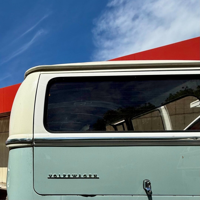 Rear of a mint green Volskwagon bus with a white upper half. In the background is a building with a red angled roof and a blue sky with wispy clouds.