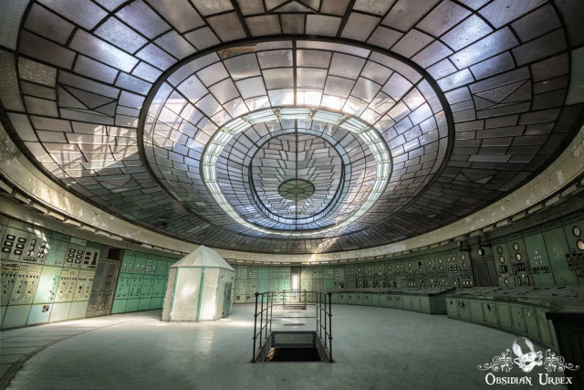 An abandoned Art Deco power plant control room featuring a grand oval glass skylight, mint-green control panels, and a central structure.