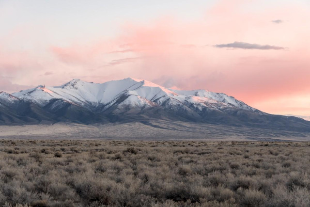 A color landscape photo of a snow capped mountain range in a flat plain of sagebrush. Wispy clouds surrounding the mountain are a light orange from the setting sun. The cloud color is also reflected in the snow facing the setting sun out of frame on the right.