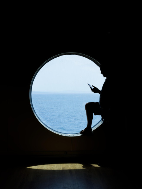 A silhouette of a person resting a foot on a circular window ledge while looking at a blue phone screen. A calm blue sea and sky are visible through the window. The sunlight shines through the window only onto a single oval spot under the window. The rest of the photo is dark. 