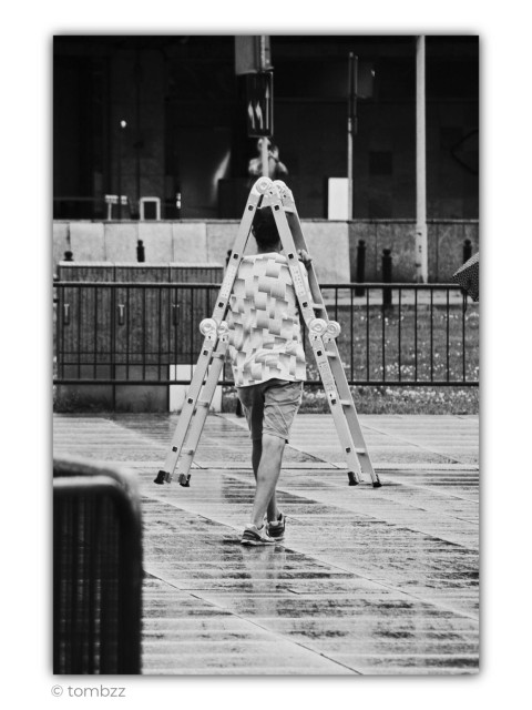 Black-and-white street photograph with a slightly surreal and humorous mood. In the center of the frame, a man walks across a wet city sidewalk carrying an unfolded aluminum ladder. The way he holds it makes the structure surround his head like an unusual geometric headdress. For a brief moment, the ladder seems less like a tool and more like an extravagant piece of avant-garde street fashion.
The man wears a light patterned T-shirt covered in interwoven rectangular shapes, knee-length shorts, and athletic shoes. Captured from behind, he appears calm and unaware of the visual joke unfolding around him. The pavement is soaked with rain, reflecting soft light and creating shimmering textures across the ground, making it clear that rain had recently passed through the city.
The background is dark and minimalist, filled with railings, street poles, and fragments of an urban crossing. On the right edge of the frame, part of another person’s umbrella quietly reinforces the rainy atmosphere. The entire scene feels like accidental street theater, where an ordinary everyday moment suddenly transforms into something playful and strangely poetic.